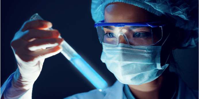 A lab technician with a safety mask and glasses looking at a beaker of blue liquid
