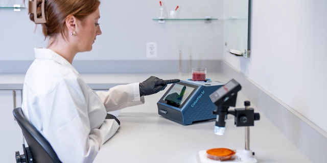 A laboratory technician examines a food sample using a benchtop spectrophotometer, with a microscope and petri dish visible in the foreground.
