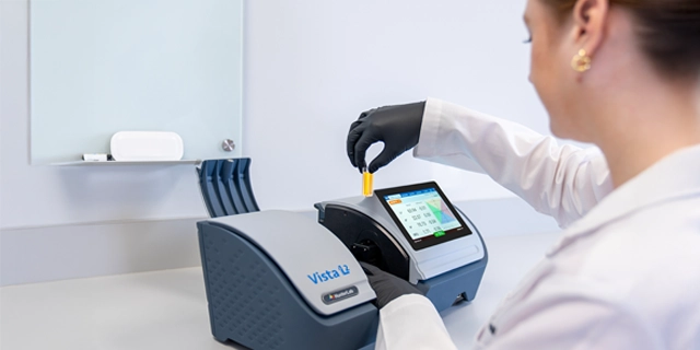 A laboratory technician places a yellow sample into a benchtop color measurement instrument, with its digital display visible.